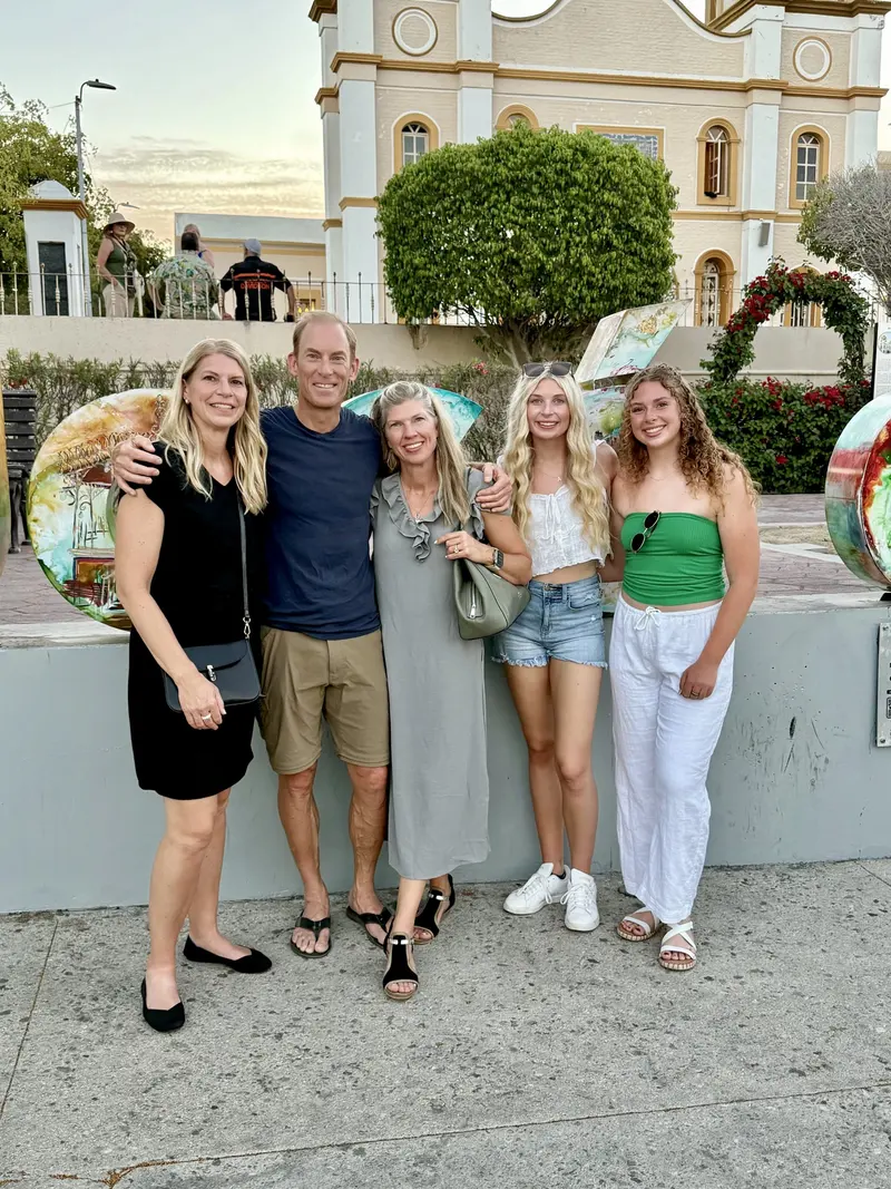 Five people stand smiling together outdoors in front of a building and art spheres; three women, one man, and one teenage girl, dressed in light, casual summer clothing.