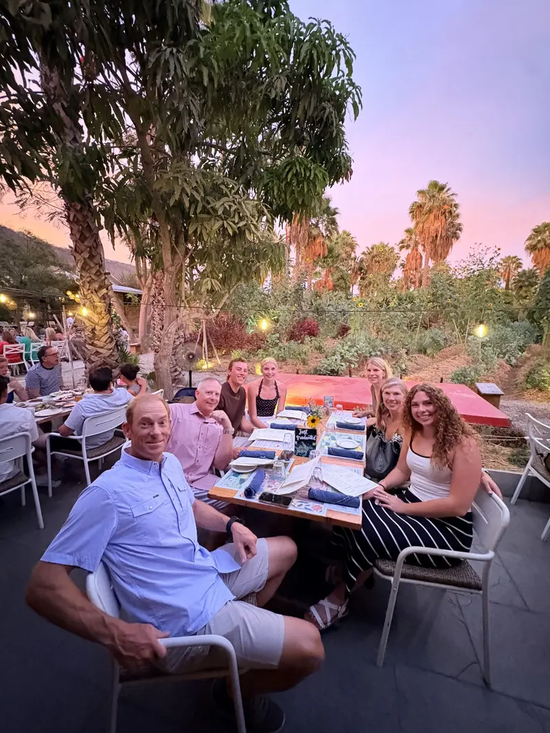 A group of seven adults sit around a table outdoors at sunset, smiling for the camera, with palm trees and greenery in the background.