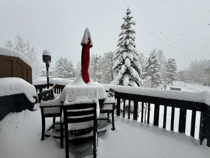 A patio table, chairs, and umbrella are covered in heavy snow on a deck. Snow falls steadily, blanketing the railing and trees in the background, creating a wintry scene.
