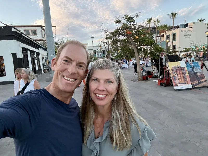 A smiling man and woman pose for a selfie outdoors in a plaza with art displays, palm trees, and people walking in the background under a blue sky with scattered clouds.