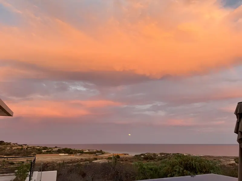 A stunning sunset sky with orange and pink clouds over a calm ocean, some buildings and vegetation in the foreground, and the moon visible near the horizon.