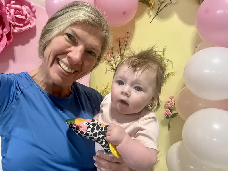 Smiling woman in a blue shirt holds a baby who is gripping a colorful toy. They are in front of pink and white balloons and pastel floral decorations.