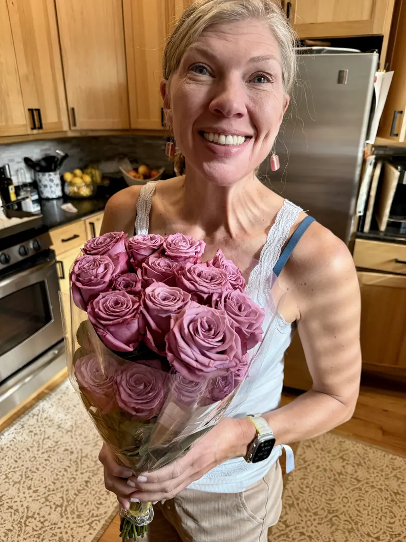Smiling woman in a kitchen holds a bouquet of mauve roses. She wears a white tank top, tan pants, and a smartwatch. Wooden cabinets, fruits, and kitchen appliances are visible in the background.