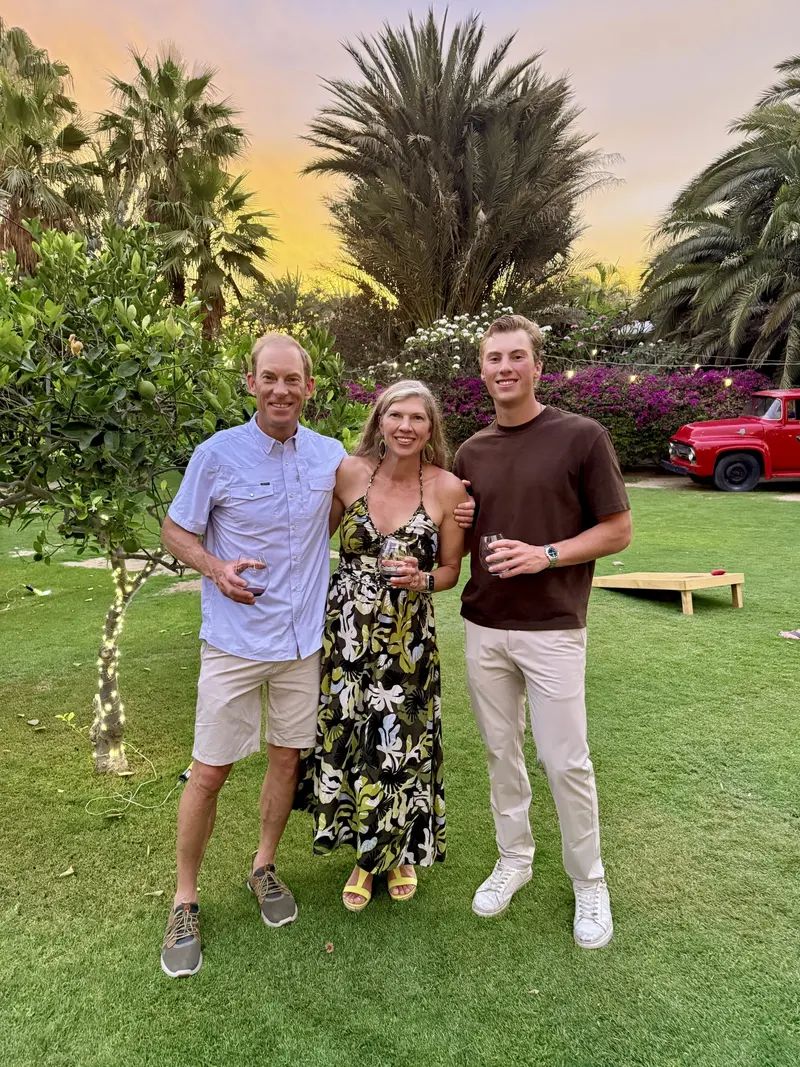 Three people stand on grass at sunset, smiling and holding drinks. Trees, flowers, a string-lit bush, and a red vehicle are in the background. A cornhole game is set up on the lawn.