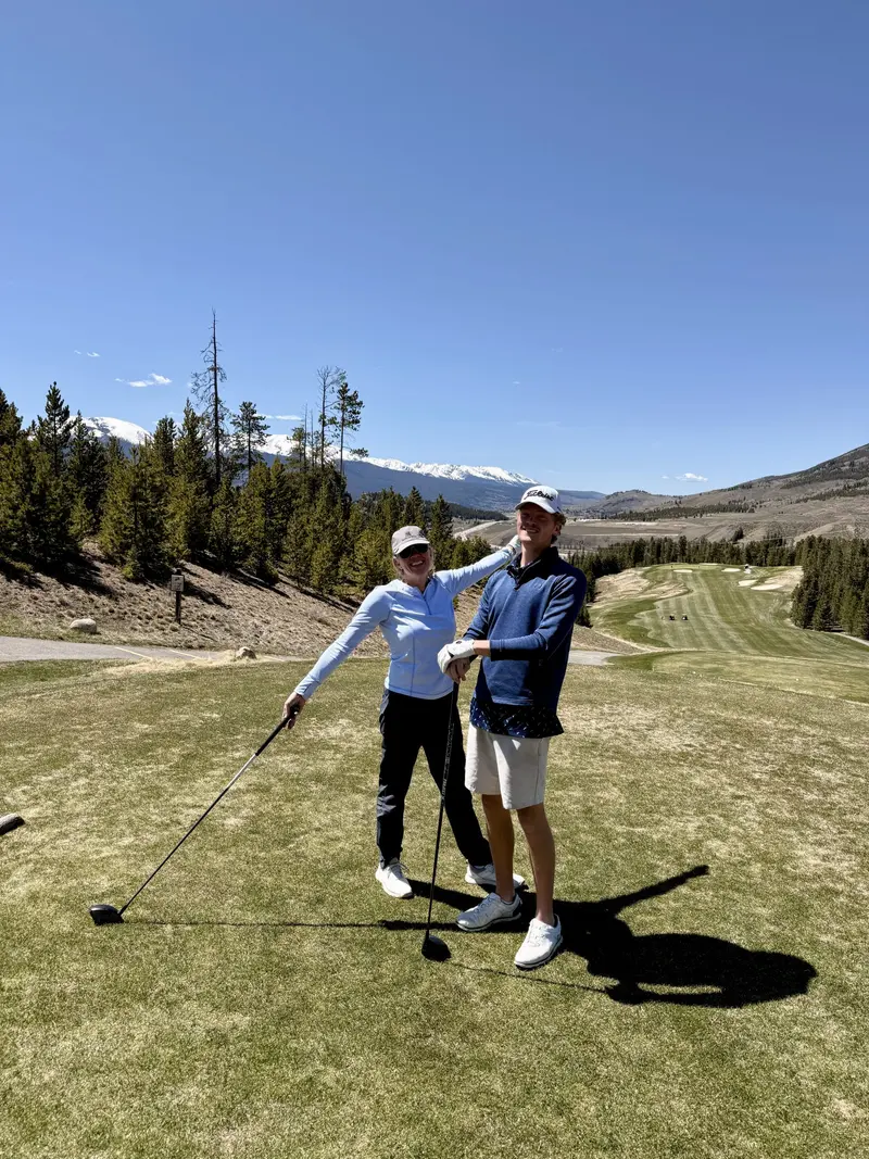 Two people in golf attire stand on a sunny golf course with mountains and trees in the background. One person playfully reaches toward the other, both smiling and holding golf clubs.