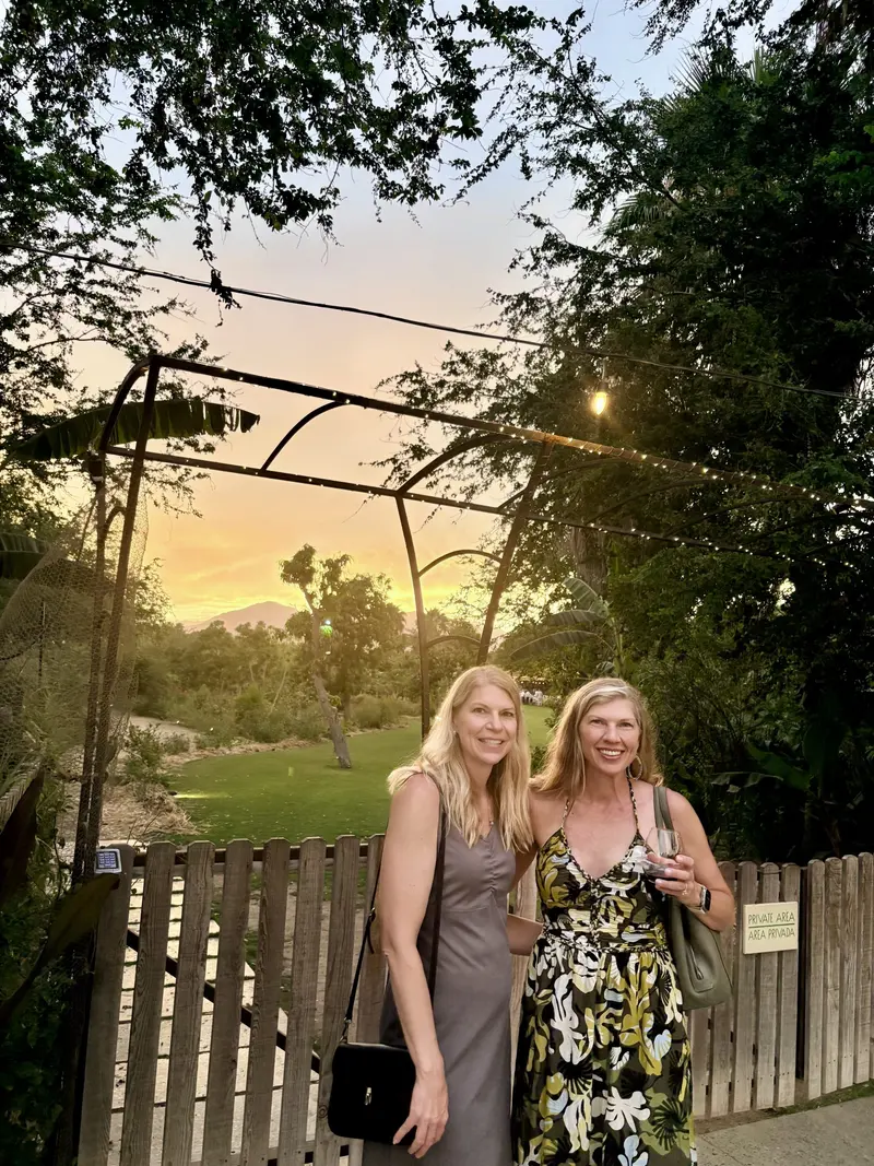 Two women smiling stand in front of a wooden gate surrounded by greenery at sunset. One wears a gray dress, the other a floral dress. The sky glows orange and pink behind them, and trees frame the scene.