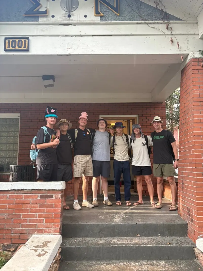 Seven young men stand smiling on a porch in front of a brick house, wearing casual clothes and hats. The house has Greek letters above the door and the number 1001. Some have backpacks, suggesting they are about to leave or have just arrived.