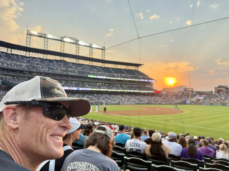 A man in sunglasses and a cap smiles at a baseball stadium crowd during sunset, with players on the field and a golden sun low in the sky.