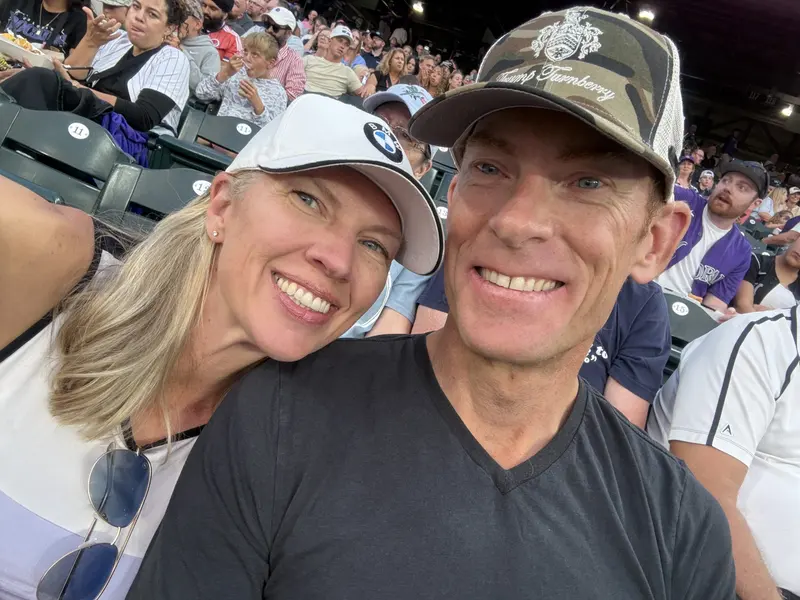 A smiling couple wearing hats takes a close-up selfie at a crowded outdoor stadium; people are seated around them, enjoying the event.