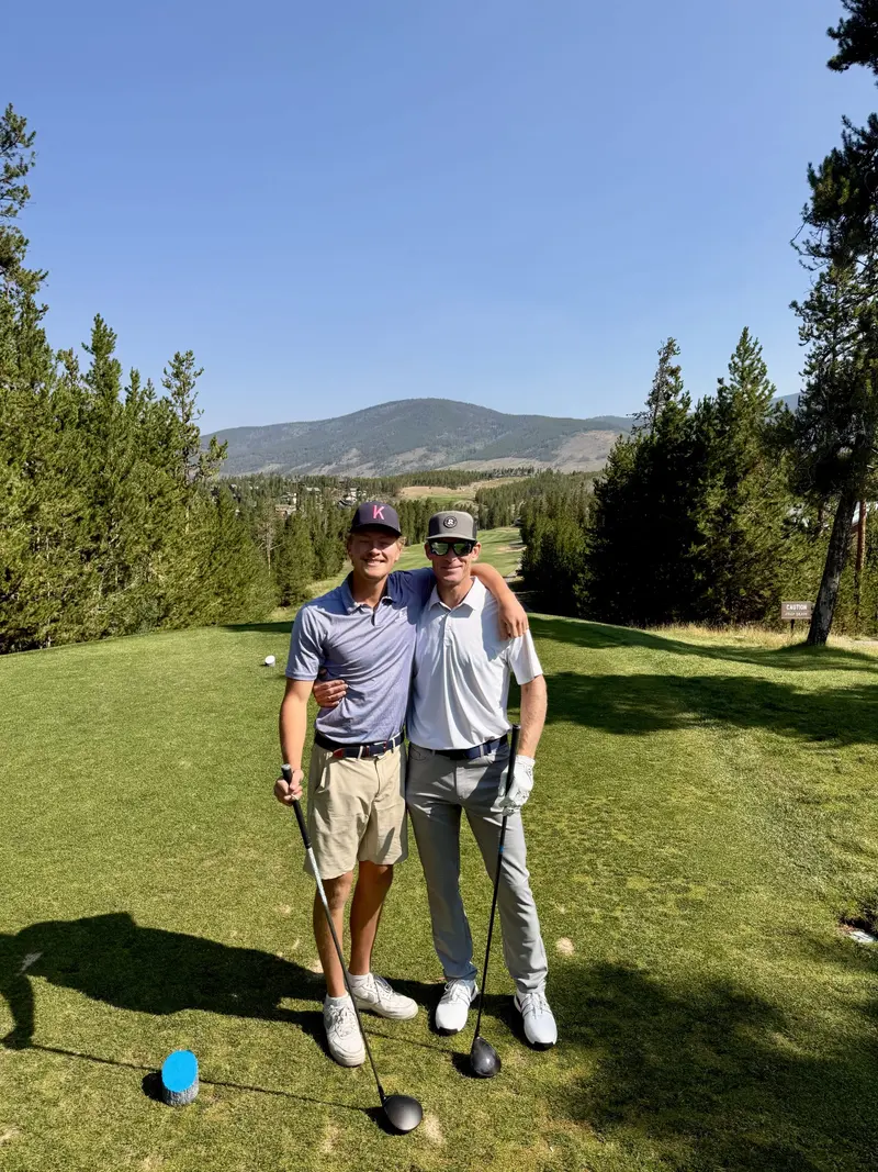 Two people pose together on a golf course tee box, each holding a golf club. Trees surround the area and mountains are visible in the background under a clear blue sky.