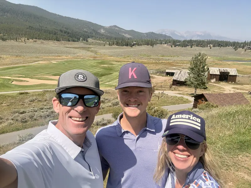 Three people in hats smiling for a selfie on a sunny day, with a golf course, rustic wooden buildings, and mountains in the background.