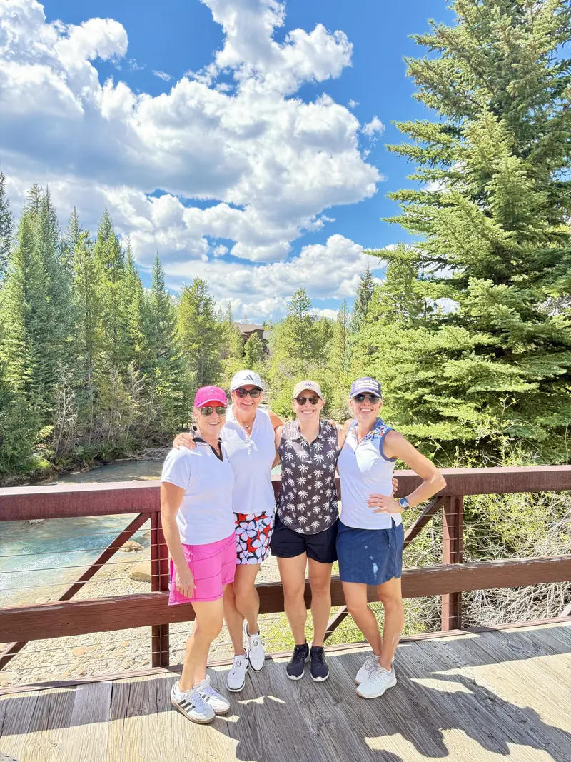 Four women stand smiling on a wooden bridge over a river, surrounded by tall green pine trees and blue sky with fluffy clouds, dressed in casual summer clothing and sunglasses.