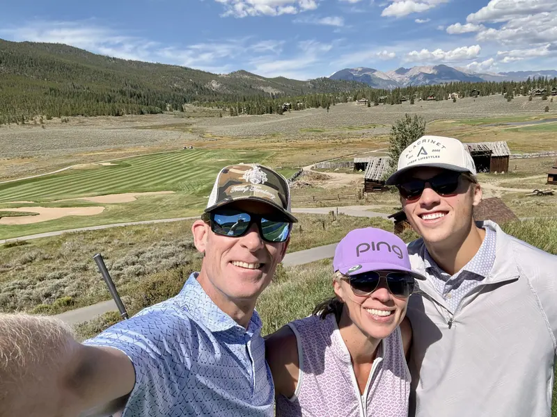 Three people wearing sunglasses and hats smile for a selfie at a golf course with rolling green hills, old wooden barns, and mountains in the background under a blue sky.