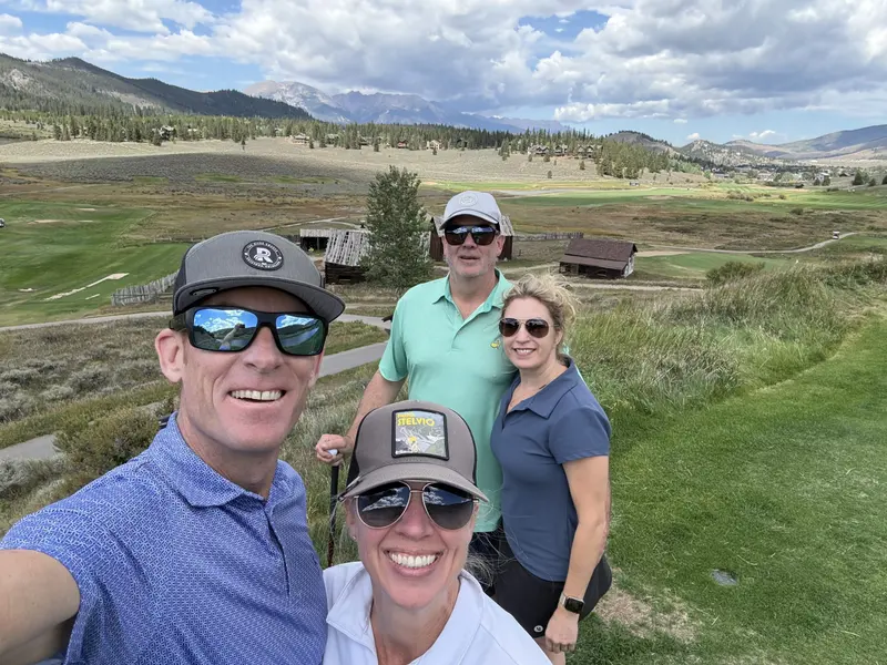 Four adults in sunglasses and hats smile on a green golf course with mountains, cabins, and clouds in the background.