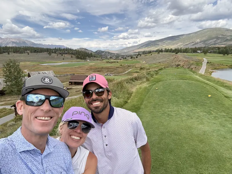 Three people wearing sunglasses and golf attire smile for a selfie on a green golf course, with mountains and cloudy skies in the background.