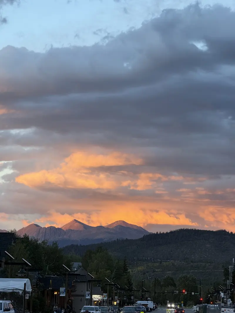 A mountain range is lit by an orange sunset under dramatic clouds, with a small town street and cars in the foreground.