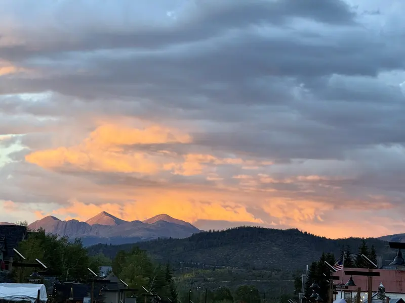 Snow-capped mountains with a glowing orange sunset sky, dramatic clouds, and forested hills in the foreground. Rooftops and trees are visible at the bottom of the image.
