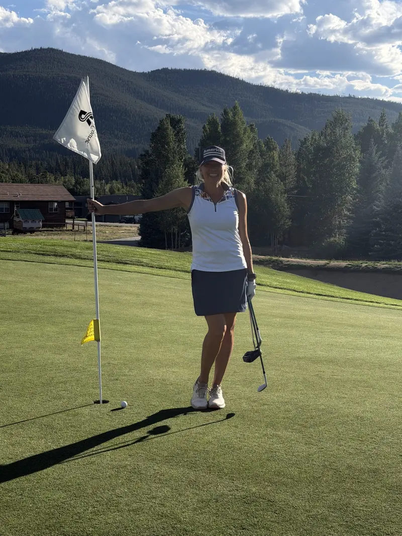 Woman in golf attire stands on a sunny green, smiling while holding a golf club and leaning on a flagstick. Trees, mountains, and a building are visible in the background.