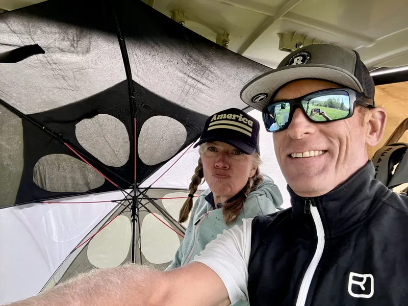 Two people sit under a large umbrella, smiling and posing for a selfie. The woman on the left wears a cap labeled America and makes a playful face. The man on the right wears sunglasses, a hat, and a black vest.