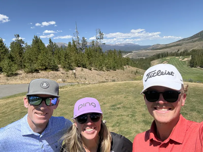 Three people wearing golf polo shirts, sunglasses, and hats smiling for a selfie on a scenic mountain golf course, with Amynakos Group.