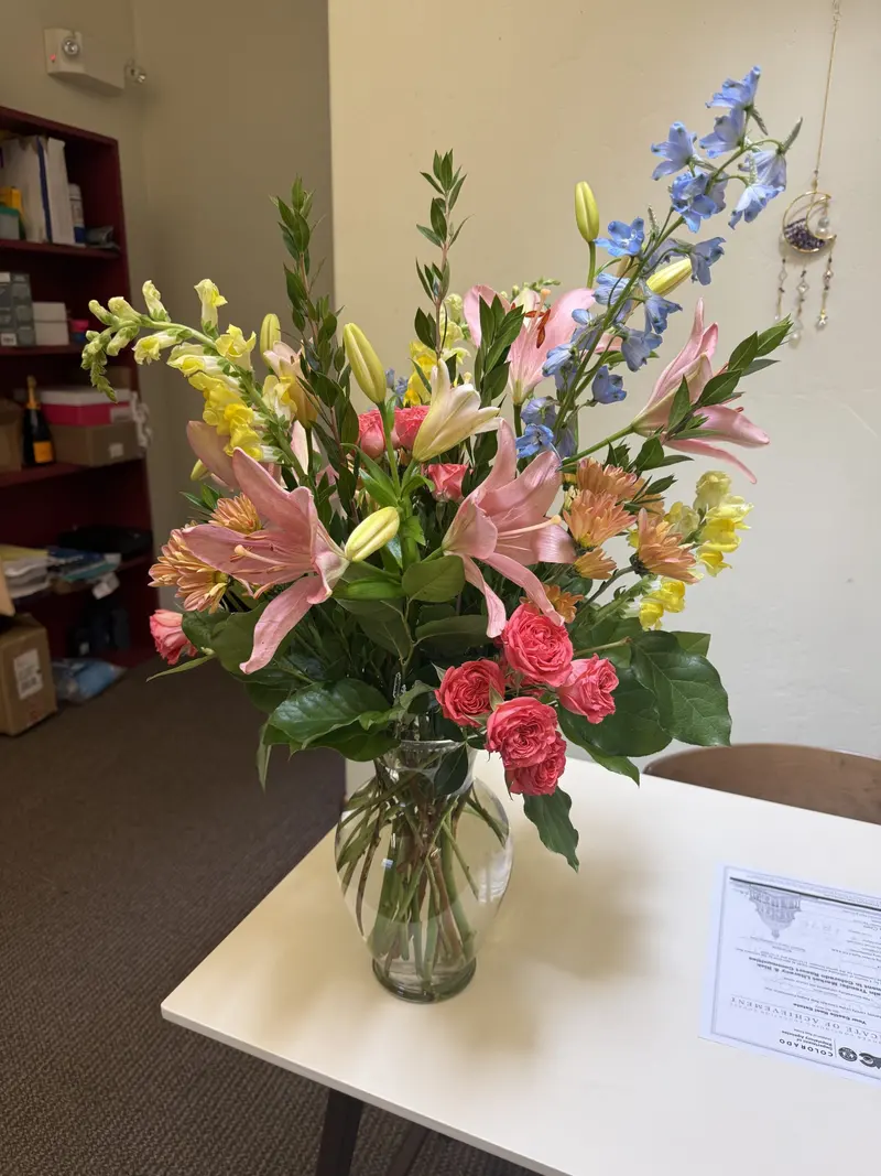Vibrant floral arrangement featuring pink lilies, blue delphiniums, and spray roses in a clear glass vase, presented by Amynakos Group.