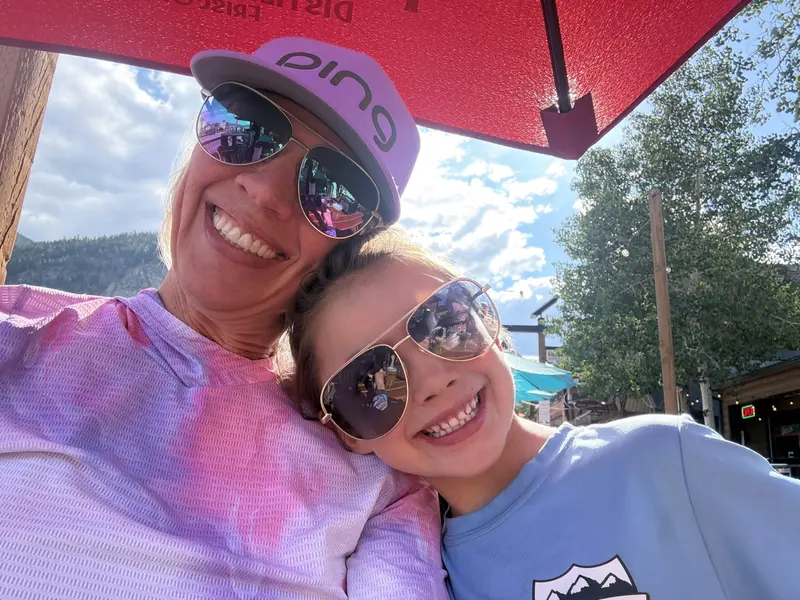 Woman and child smiling together under an outdoor restaurant umbrella in a mountain town setting, presented by Amynakos Group