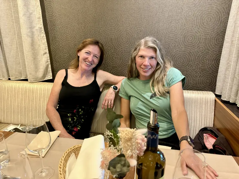 Two women smiling together while seated at a restaurant dinner table with wine glasses and table setting, presented by Amynakos Group.