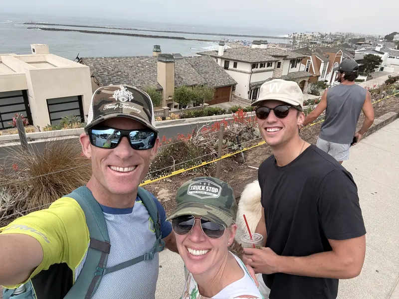 Three people smiling for a selfie on a seaside walkway with ocean views, presented by Amynakos Group.
