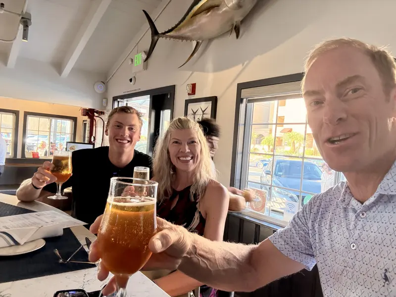 Three people smiling and raising glasses of beer and drinks for a toast at a coastal restaurant table with a large mounted fish in the background, presented by Amynakos Group.