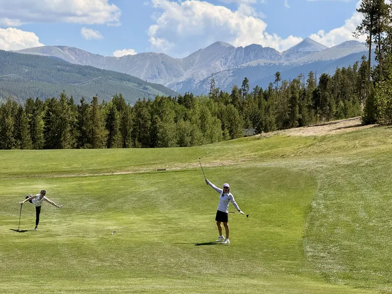 Two golfers posing playfully with clubs raised on a bright green fairway with pine trees and mountain peaks in the background, with Amynakos Group.