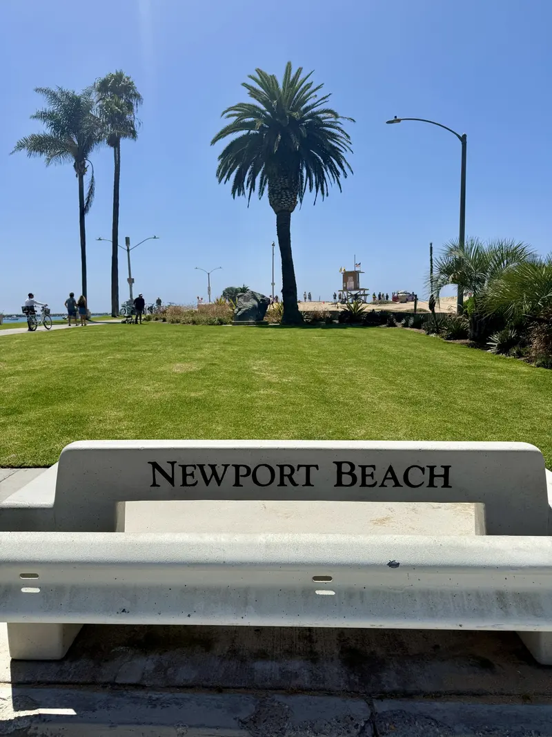 Concrete bench labeled Newport Beach overlooking a grassy park with palm trees under a bright blue sky, with Amynakos Group.