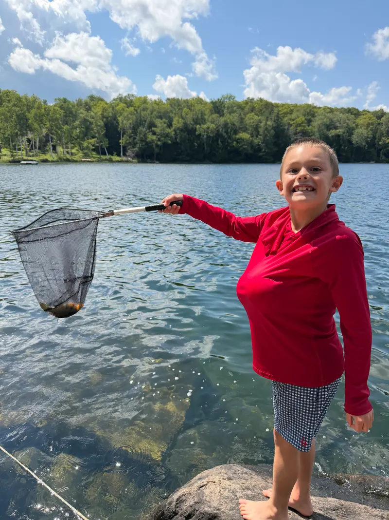 A smiling child in a red shirt stands on a rock by a lake, holding a fishing net containing a small fish.
