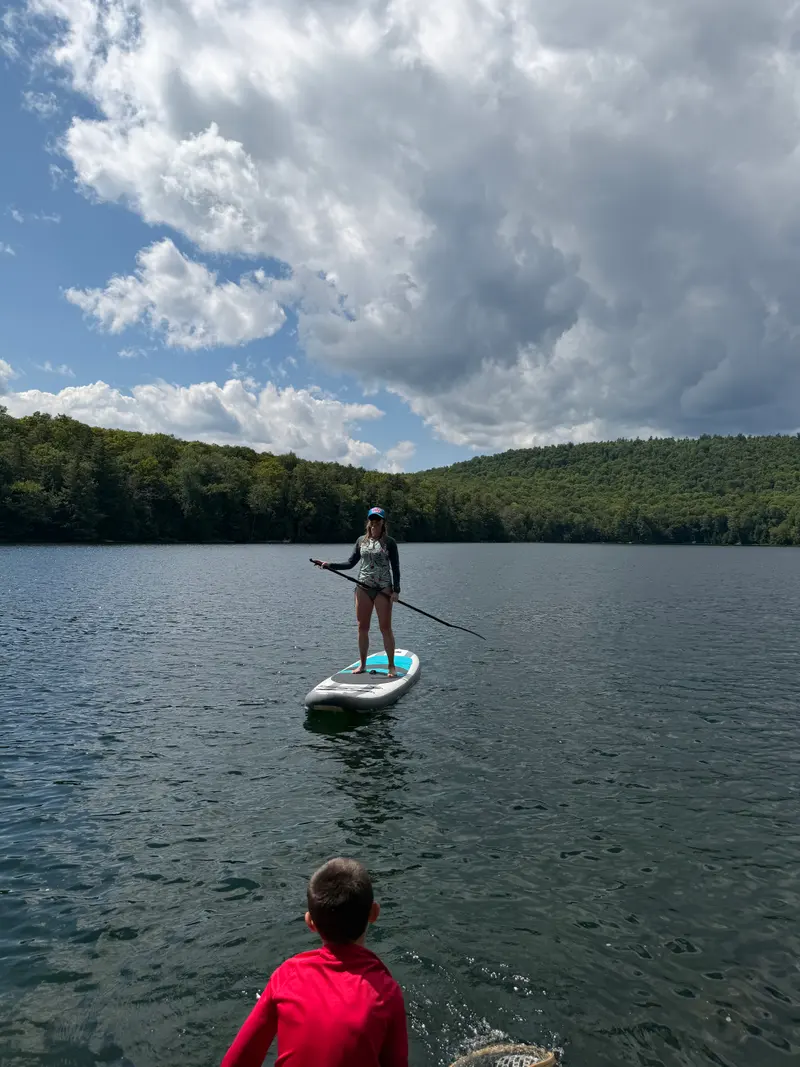 A person stands on a paddleboard on a lake while another person looks on from the foreground.