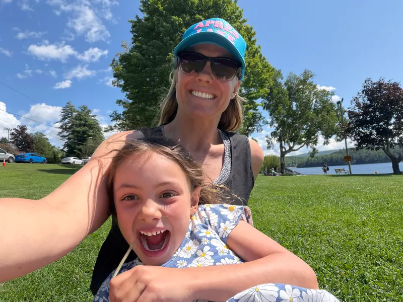 A woman wearing a blue baseball cap and sunglasses takes a selfie while holding a laughing child on a grassy field by a lake.