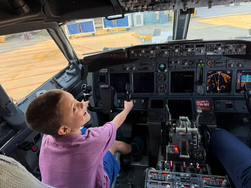 A young boy sits in the pilots seat of an airplane cockpit, holding the control yoke and looking out the window.