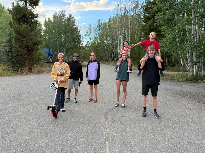 A family of six stands together on a paved road lined with trees, with one child riding on anothers shoulders and a boy holding a scooter in the foreground.
