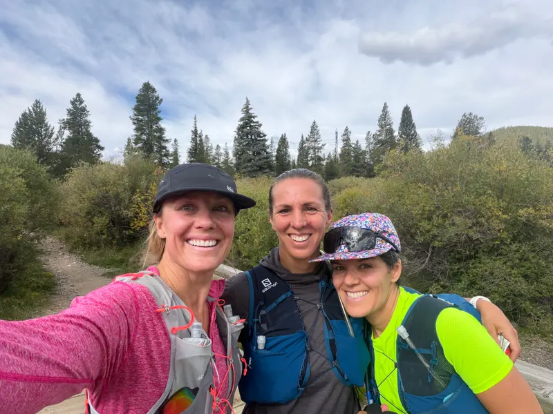 Three women in athletic wear and running vests pose for a selfie on a trail surrounded by trees.