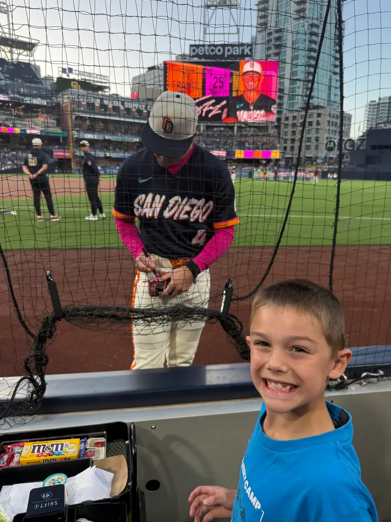 A young boy smiles for the camera at a baseball stadium while a player signs an autograph in the background.