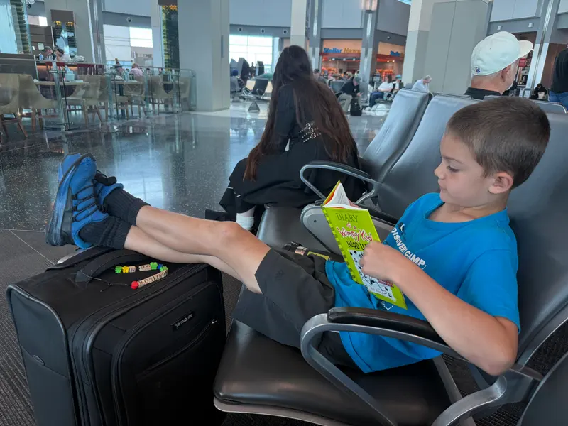 A boy in a blue t-shirt sits in an airport gate area reading a Diary of a Wimpy Kid book with his feet propped up on a black carry-on suitcase.