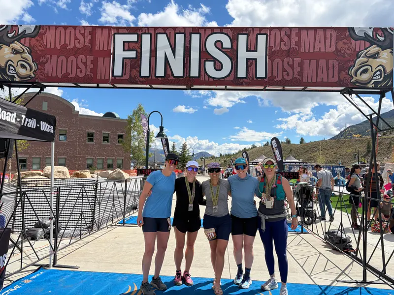 Five women wearing race bibs and medals stand smiling under a large FINISH arch at an outdoor race event.
