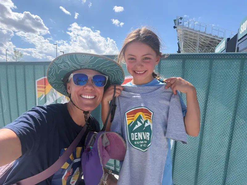 A woman wearing a sun hat and sunglasses takes a selfie with a young girl who is holding up a gray Denver Summit FC soccer t-shirt.