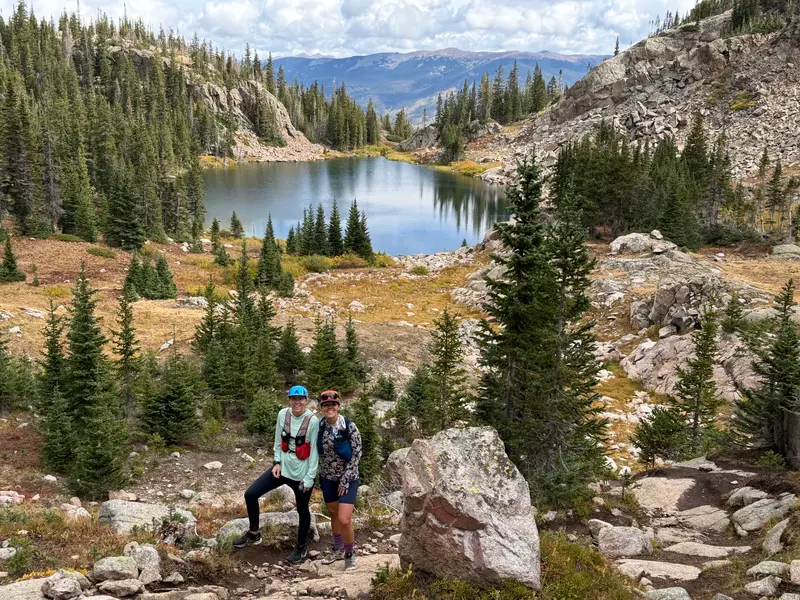 Two hikers stand on a mountain trail overlooking a lake surrounded by pine trees and rocky slopes.