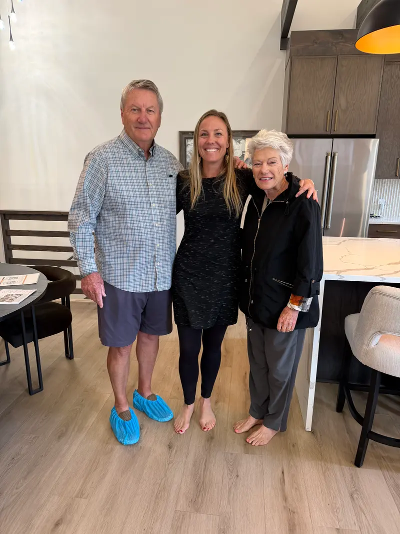 An older man in a plaid shirt and blue shoe covers, a middle-aged woman in a black dress, and an older woman in a black jacket stand side-by-side in a kitchen.