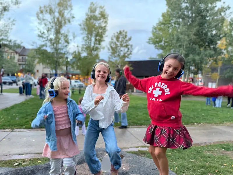 Three girls wearing headphones dance together on a sidewalk in a park.