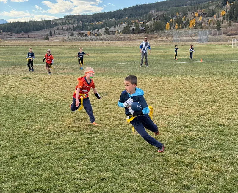 A young boy in a blue hoodie carries a football across a grassy field during a flag football game, with another player chasing him and a referee nearby.