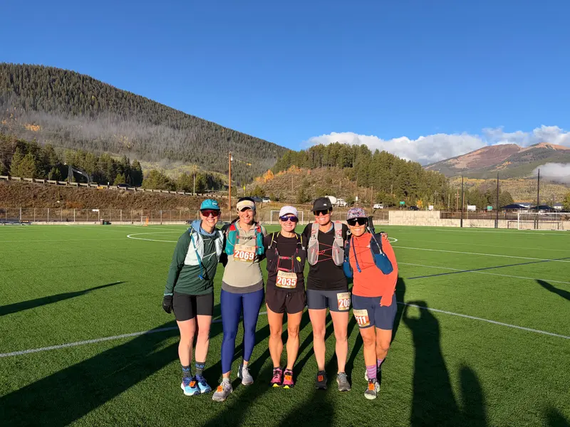 Five women wearing running gear and race bibs stand together on a turf athletic field with mountains in the background.